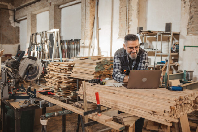 Carpenter in wood workshop using laptop and working on project, online shopping or banking.